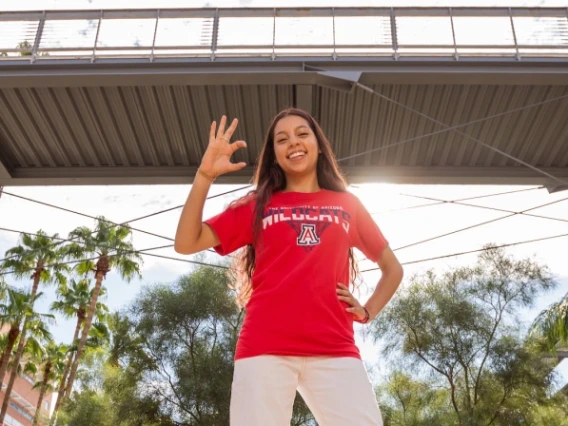 Emily Aguilar stands on a concrete wall making the wildcat hand sign (pinky, ring and middle fingers making a "w" and thumb and pointer fingers curved to make a "c") with the Aerospace and Mechanical Engineering courtyard and bridge in the background.