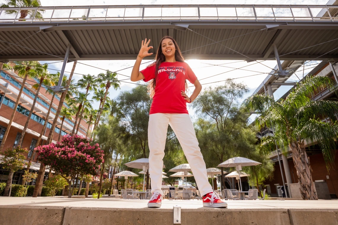 Emily Aguilar stands on a concrete wall making the wildcat hand sign (pinky, ring and middle fingers making a "w" and thumb and pointer fingers curved to make a "c") with the Aerospace and Mechanical Engineering courtyard and bridge in the background.