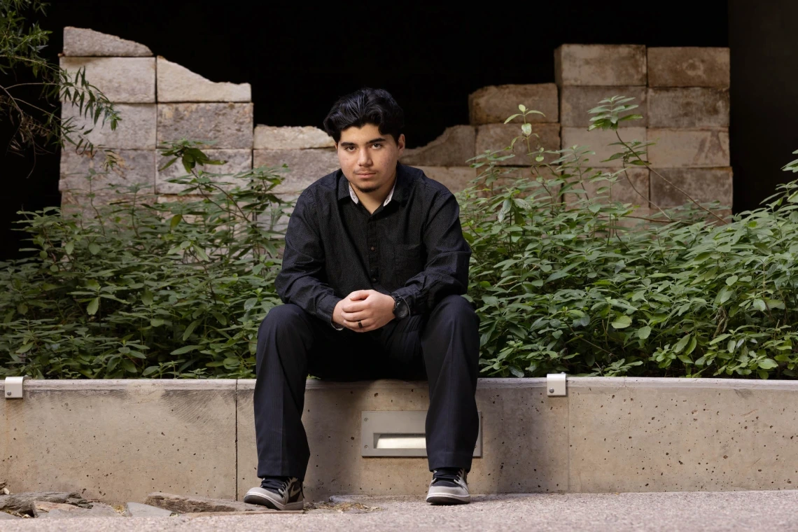 Marcelino Perez seated, wearing all black, on a concrete wall.